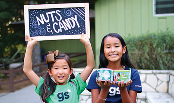 Girl holding up sign that says "Nuts & Candy" and the other girl is holding snack packages
