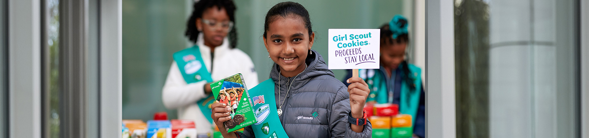  girl scouts selling cookies with one girl in front of booth holding sign that says "girl scout cookie proceeds stay local" 