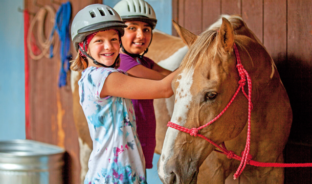 two preteen girls in a stable smiling while petting a brown horse