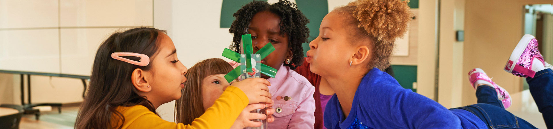  four little girls of different ethnicities leaning over a table working on their propeller science experiment 