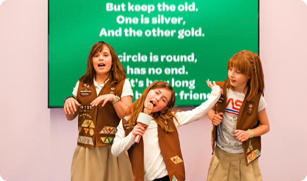 Three girls singing into a microphone in front of a television that is displaying song lyrics