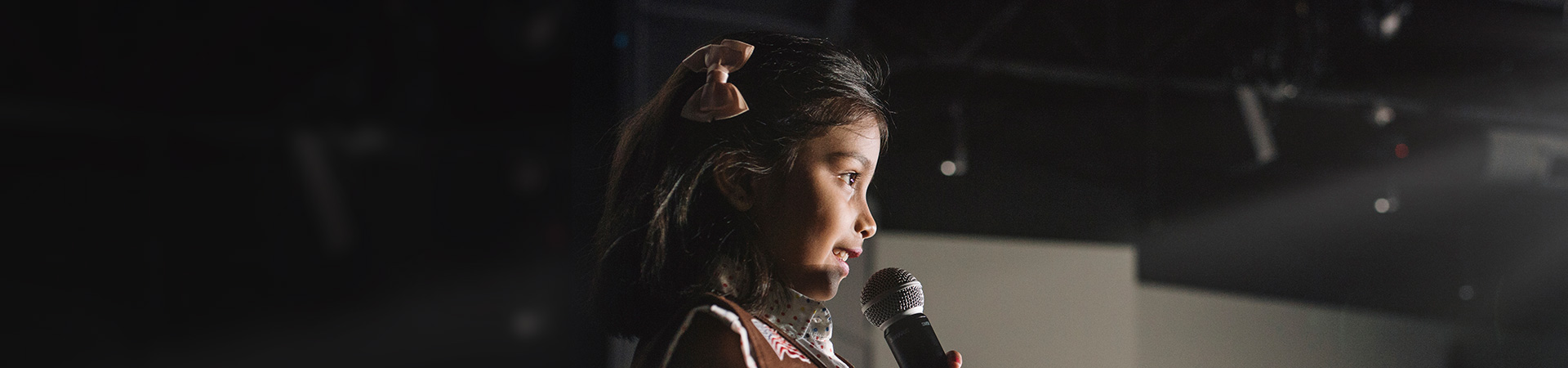  a cute little girl in a brown girl scout vest speaking into a microphone in a dark theatre 