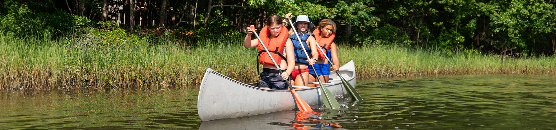  three older girls wearing life vests in a canoe on water rowing with their oars 