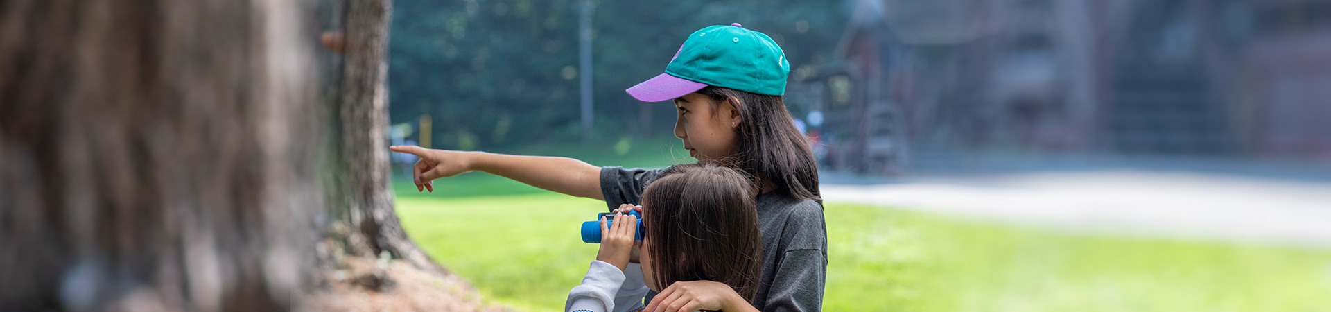  two young girls exploring the outdoors; the older girl is pointing while the younger girl is using binoculars 